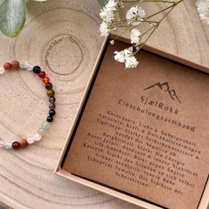 Könnte beinhalten: Ein mehrfarbiges Perlenarmband mit verschiedenen Edelsteinen, darunter rote, orangefarbene, grüne und schwarze Perlen. Das Armband wird auf einer Holzoberfläche neben einer Schachtel mit deutschem Text präsentiert. Weiße Blumen und grüne Blätter sind im Hintergrund.