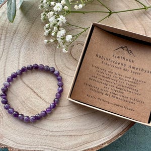 Könnte beinhalten: Ein violettes Amethyst-Armband mit einer braunen Schachtel, auf der "SjælRokk Rubinfarbene Amethyst Schutzstein der Seele" steht und die Vorteile von Amethyst beschreibt.