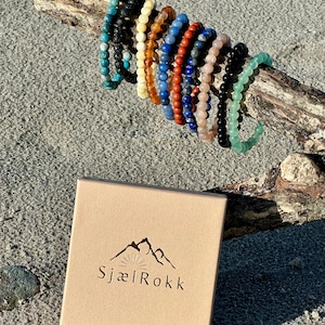 K&ouml;nnte beinhalten: Ein Set aus 10 bunten Perlenarmb&auml;ndern, jedes mit einem anderen Stein, auf einem St&uuml;ck Treibholz pr&auml;sentiert. Die Armb&auml;nder sind in einem Halbkreis angeordnet, wobei sich die Enden der Armb&auml;nder &uuml;berlappen. Die Armb&auml;nder sind aus nat&uuml;rlichen Steinen gefertigt, darunter Lavastein, Bernstein, roter Jaspis, Lapislazuli, Rosenquarz, schwarzer Onyx und gr&uuml;ner Aventurin. Die Armb&auml;nder sind alle etwa 18 cm lang. Die Armb&auml;nder sind in einer kleinen Pappbox mit dem Logo "Sjael Rokk" auf der Oberseite verpackt.