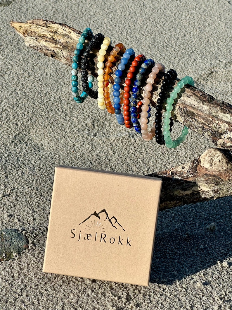 K&ouml;nnte beinhalten: Ein Set aus 10 bunten Perlenarmb&auml;ndern, jedes mit einem anderen Stein, auf einem St&uuml;ck Treibholz pr&auml;sentiert. Die Armb&auml;nder sind in einem Halbkreis angeordnet, wobei sich die Enden der Armb&auml;nder &uuml;berlappen. Die Armb&auml;nder sind aus nat&uuml;rlichen Steinen gefertigt, darunter Lavastein, Bernstein, roter Jaspis, Lapislazuli, Rosenquarz, schwarzer Onyx und gr&uuml;ner Aventurin. Die Armb&auml;nder sind alle etwa 18 cm lang. Die Armb&auml;nder sind in einer kleinen Pappbox mit dem Logo "Sjael Rokk" auf der Oberseite verpackt.