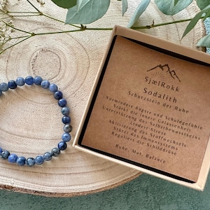 Könnte beinhalten: Ein blaues Sodalith-Armband aus Edelsteinen mit einer kleinen Schachtel, die eine Beschreibung der Eigenschaften des Steins enthält. Die Schachtel trägt die Aufschrift "SjælRokk Sodalith Schutzstein der Ruhe" und listet die Vorteile des Tragens von Sodalith auf, darunter die Reduzierung von Angstzuständen, die Förderung von innerem Frieden und die Unterstützung des Selbstbewusstseins.
