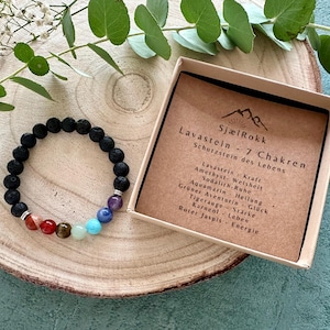 Könnte beinhalten: Ein schwarzes Lavastein-Armband mit farbigen Chakra-Perlen, präsentiert auf einer Holzscheibe mit einer Box mit deutschem Text. Das Armband hat rote, orange, gelbe, grüne, blaue und violette Perlen.