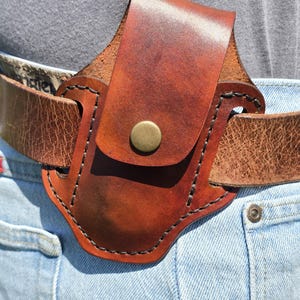 May include: A brown leather belt holster with a flap secured by a brass button. The holster is attached to a brown leather belt worn over blue jeans. The leather has a distressed appearance.