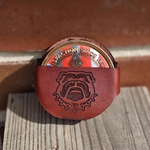 May include: A brown leather holder featuring an embossed bulldog face design on the front. Inside the holder sits a circular metal coin with a red and white striped background, a central W logo, and the text SATISFACTION SINCE 1825 printed on it.