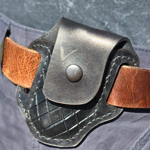 May include: A black leather belt pouch featuring a basket-weave embossed pattern on the lower section and a single metal snap closure. The pouch is threaded onto a wide, textured brown leather belt, which is worn over dark gray fabric trousers.