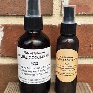 May include: Two dark brown glass bottles of natural cooling mist. The larger bottle is labeled 4oz, the smaller 2oz. Both have black spray tops and white labels with product information. The background is a brick wall.