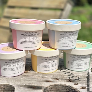 May include: A selection of natural odor absorbers in various colours. Each container has a white base and lid, with labels indicating scents such as "Cozy Sweet Chur" and "Cool Fresh Air." The containers are displayed on a wooden surface.