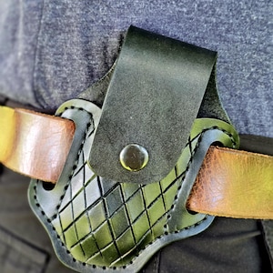 May include: Black leather holster with a diamond pattern, secured to a brown leather belt. The holster features a flap with a metal snap closure. The design is visible against a gray shirt and black pants.
