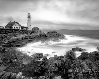 May include: A black and white photograph of a lighthouse on a rocky coast with crashing waves. The lighthouse is tall and white with a black roof. The waves are crashing against the rocks in the foreground, creating a sense of motion and energy.