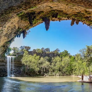 May include: A view of a waterfall cascading into a pool of water, framed by a natural rock archway. The water is clear and blue, and the surrounding trees are green. The sky is a bright blue.