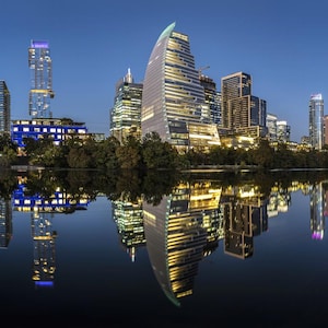May include: A panoramic view of the Austin skyline at dusk, with several skyscrapers reflected in the still water of a lake. The tallest building is a modern glass and steel structure with a distinctive curved design.