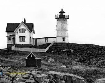 May include: A black and white photograph of a lighthouse and a house on a rocky cliff. The lighthouse is tall and white with a black roof and a small window. The house is a two-story building with a white exterior and a black roof.