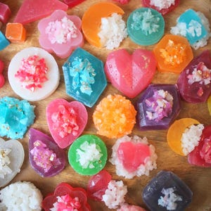 Könnte beinhalten: Sortiment aus bunten, handgefertigten Seifen in verschiedenen Formen, darunter Herzen, Blumen und geometrische Designs. Jede Seife ist mit weißen oder rosa Zuckerkristallen verziert.