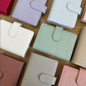 Op de afbeelding: Een verzameling kleine, getextureerde notitieboekjes in verschillende pastelkleuren, waaronder roze, lavendel en mintgroen. Elk notitieboekje heeft een bijpassende tabsluiting.