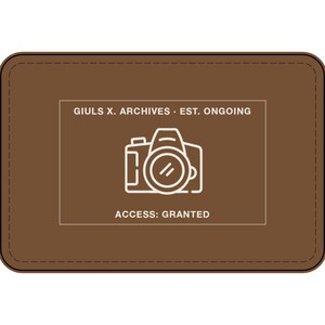 May include: A rectangular brown leather patch with rounded corners and stitched edges. The center features a white line-art graphic of a camera, with the text GIULS X. ARCHIVES EST. ONGOING above and the text ACCESS: GRANTED below the camera.
