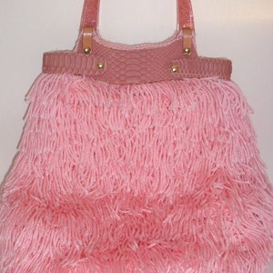 Puede incluir: Un bolso de mano rosa con flecos largos. El bolso tiene un material rosa similar a la piel de serpiente en la parte superior y asas de plástico transparente.