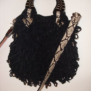 Op de afbeelding: Zwarte handtas met een franje-ontwerp en een slangleren riem. De handtas heeft een houten handvat met een geometrisch patroon.