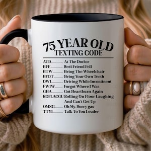 May include: White ceramic mug with a black interior and handle. The mug features the text "75 YEAR OLD TEXTING CODE" with a list of texting acronyms and their meanings, such as "ATD... At The Doctor" and "BFF... Best Friend Fell."