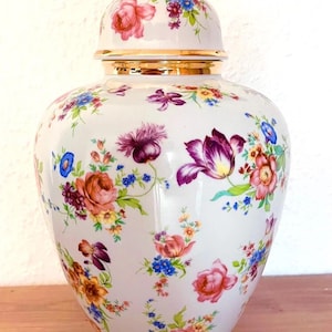 Puede incluir: Un jarrón de cerámica blanco con tapa, adornado con un vibrante estampado floral en tonos rosa, morado, naranja y azul. La tapa y la base tienen ribetes dorados. Una pieza decorativa para el hogar.