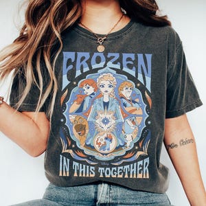 May include: Dark gray t-shirt featuring the word "FROZEN" in blue, with a graphic of characters from the movie. The phrase "IN THIS TOGETHER" is below the graphic. The design includes Elsa, Anna, Kristoff, Olaf, and Sven.