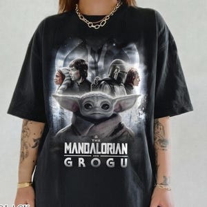 May include: Black oversized t-shirt featuring a graphic of characters from The Mandalorian series, including Grogu. The shirt has the text "THE MANDALORIAN AND GROGU" in white. The words "BLACK COMFORT COLORS" are printed at the bottom.