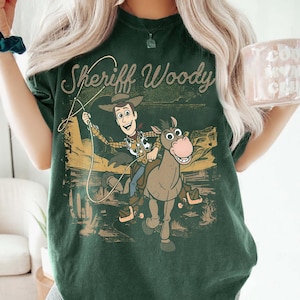 May include: Dark green t-shirt featuring a graphic of Sheriff Woody riding a horse with the text "Sheriff Woody" in a vintage style. The design includes a cowboy hat, rope, and desert landscape.