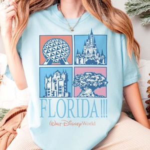 May include: Light blue t-shirt featuring four square graphics with iconic Florida landmarks. The shirt includes the text "FLORIDA!!!" and "Walt Disney World" in a playful font. The shirt is a comfortable, casual style.