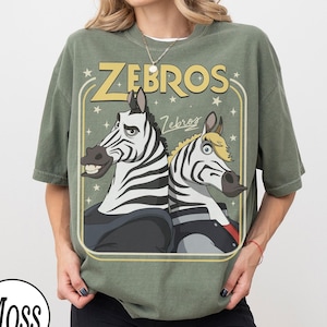 May include: A moss green t-shirt featuring a cartoon graphic of two zebras with the text "ZEBROS" in yellow. The zebras are wearing jackets and have expressive faces. The shirt has a vintage, slightly faded look.