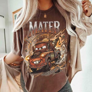 Pode incluir: Camiseta marrom com um desenho do Mate do filme Carros. O design inclui a palavra "MATER" acima do personagem, com raios e elementos mec&acirc;nicos. A camiseta tem um corte folgado.
