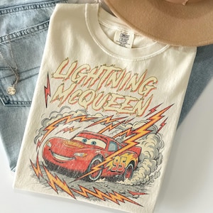 May include: Cream-colored t-shirt featuring a graphic of Lightning McQueen from the Cars movie. The design includes the text "Lightning McQueen" in red and yellow, along with a red race car and the number 95. The shirt is paired with blue jeans and a necklace.