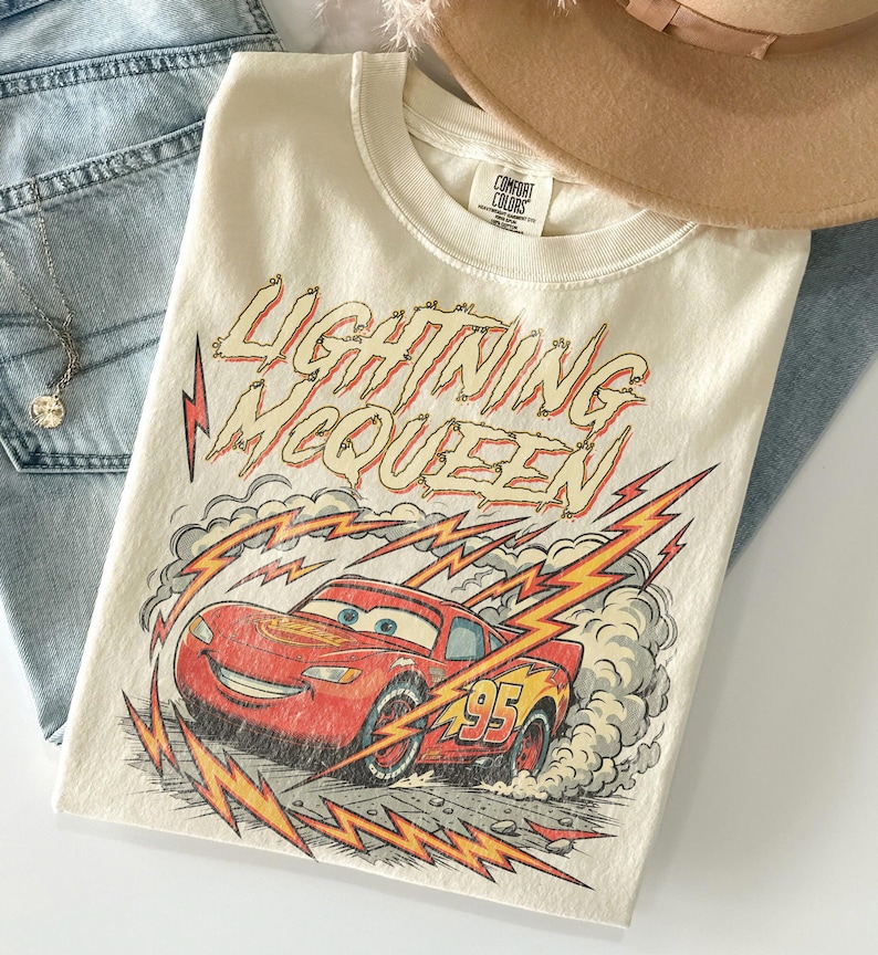 May include: Cream-colored t-shirt featuring a graphic of Lightning McQueen from the Cars movie. The design includes the text "Lightning McQueen" in red and yellow, along with a red race car and the number 95. The shirt is paired with blue jeans and a necklace.