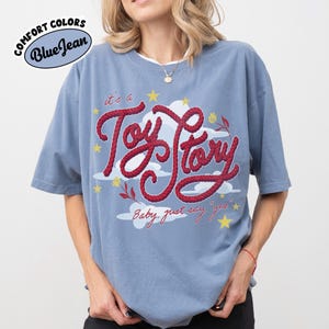 May include: A blue jean colored t-shirt featuring a graphic print with the red rope-style text "it's a Toy Story" and "Baby, just say yes". The design includes white clouds, yellow stars, and small red flowers. The shirt has a crew neckline.