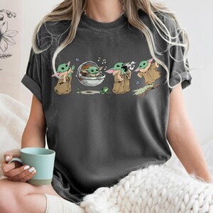 May include: Dark grey t-shirt with a cartoon illustration of a character from a popular science fiction series. The graphic shows the character in various poses, with musical notes and a frog. The character is wearing a brown cloak.