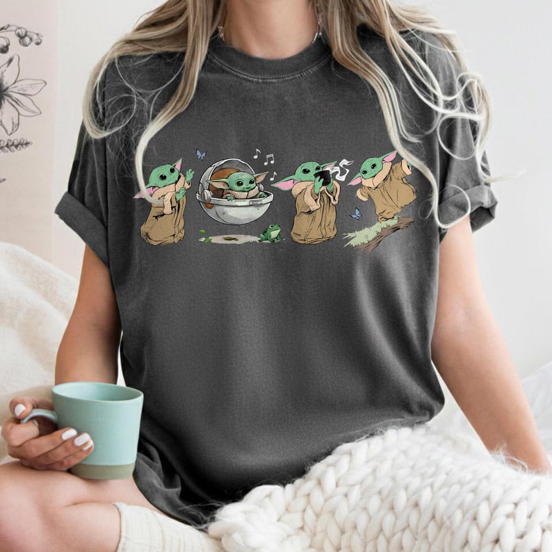 May include: Dark grey t-shirt with a cartoon illustration of a character from a popular science fiction series. The graphic shows the character in various poses, with musical notes and a frog. The character is wearing a brown cloak.
