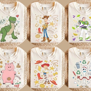 May include: Six off-white t-shirts with colorful cartoon character designs. The designs include a green dinosaur, Woody, Buzz Lightyear, Hamm the pig, Jessie, and Forky. Each shirt is folded and displayed on a woven surface.