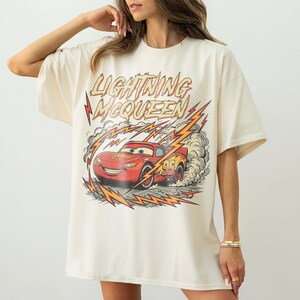 May include: Cream-colored t-shirt featuring a graphic of Lightning McQueen, a red race car, with the text "Lightning McQueen." The design includes lightning bolts and smoke effects, creating a dynamic and energetic look. The shirt has short sleeves and a relaxed fit.