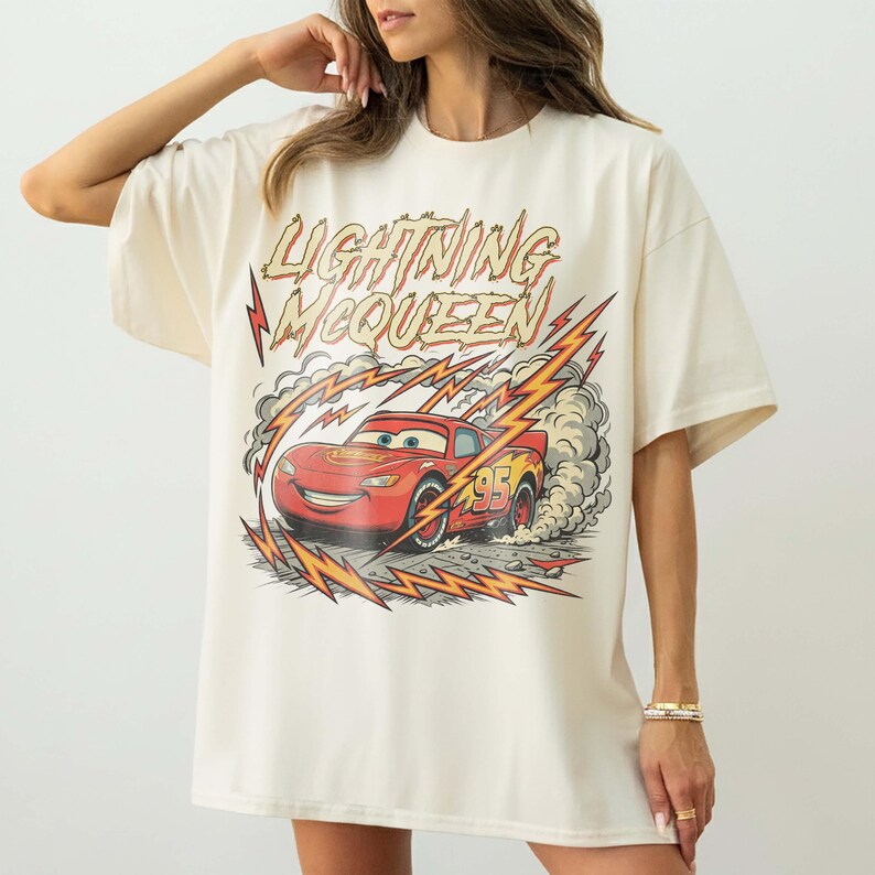 May include: Cream-colored t-shirt featuring a graphic of Lightning McQueen, a red race car, with the text "Lightning McQueen." The design includes lightning bolts and smoke effects, creating a dynamic and energetic look. The shirt has short sleeves and a relaxed fit.