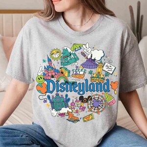May include: A gray t-shirt featuring a colorful Disneyland graphic. The design includes iconic park elements like a castle, a Ferris wheel, and the Disneyland logo. The shirt is a casual, round-neck style.