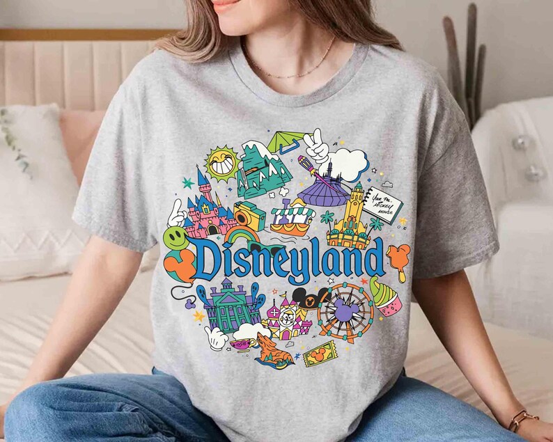 May include: A gray t-shirt featuring a colorful Disneyland graphic. The design includes iconic park elements like a castle, a Ferris wheel, and the Disneyland logo. The shirt is a casual, round-neck style.