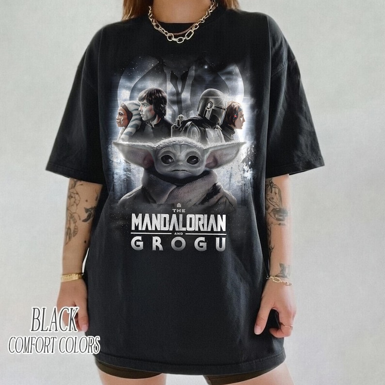 May include: Black oversized t-shirt featuring a graphic of characters from The Mandalorian series, including Grogu. The shirt has the text "THE MANDALORIAN AND GROGU" in white. The words "BLACK COMFORT COLORS" are printed at the bottom.