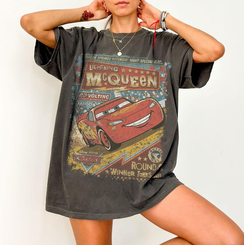 Pu&ograve; includere: T-shirt grigio scuro con Lightning McQueen del film Disney Pixar Cars. La grafica include il testo "Lightning McQueen" e "Winner Takes All" in stile vintage. La maglietta ha maniche corte e una vestibilit&agrave; ampia.