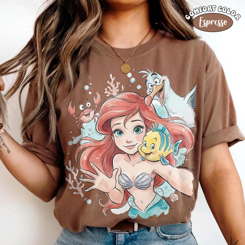 Op de afbeelding: Een espresso-kleurig t-shirt met een grafische print van een zeemeermin met rood haar, een gele en blauwe vis, een kleine krab en een witte meeuw. Het ontwerp bevat koraal en bubbels, met de tekst Comfort Color Espresso in de hoek.