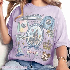 Pode incluir: Camiseta cor de orquídea com um design lúdico com um castelo, trem, suéteres, chaves e um biscoito do Mickey Mouse. A camisa inclui uma faixa que diz "Most Magical Place".