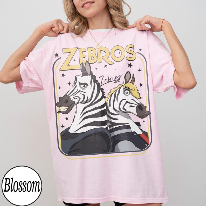 May include: Pink t-shirt featuring a cartoon graphic of two zebras with the text "Zebros" in a retro style. The zebras have black and white stripes and are wearing outfits. The shirt has short sleeves and a round neckline.
