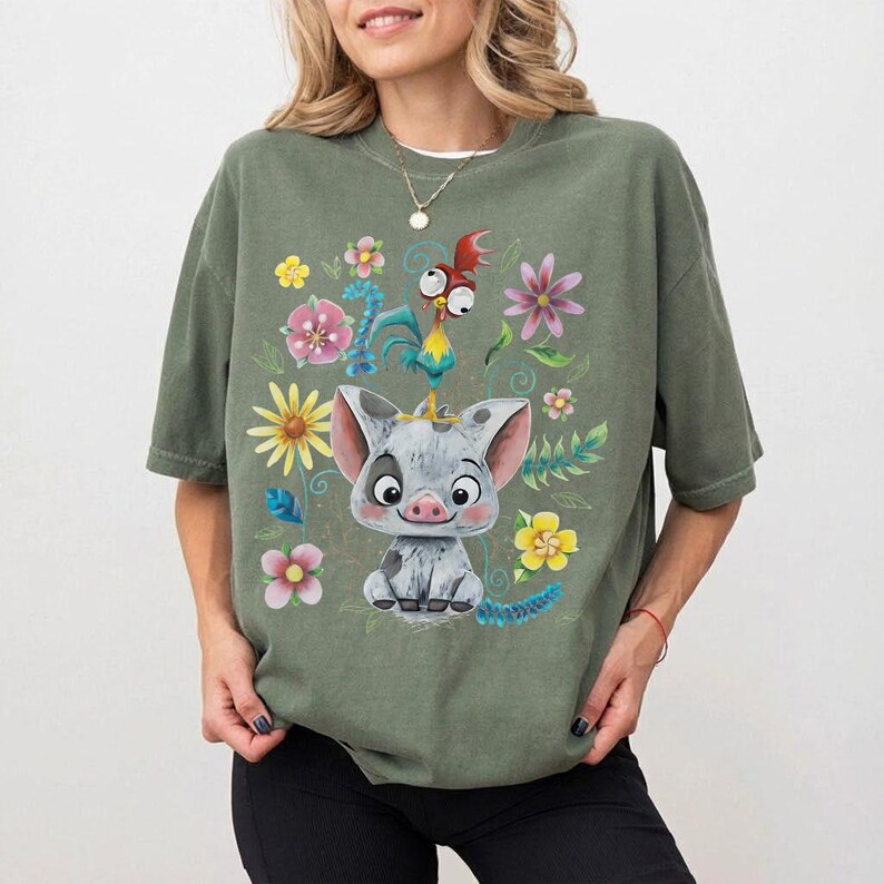 K&ouml;nnte beinhalten: Olivgr&uuml;nes T-Shirt mit einem Cartoon-Schwein und Hahn-Design, umgeben von bunten Blumen und Bl&auml;ttern. Das Schwein ist grau und wei&szlig;, und der Hahn ist rot, gelb und blau. Das Shirt hat kurze &Auml;rmel und einen Rundhalsausschnitt.