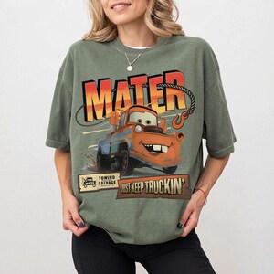 以下が含まれることがあります： 映画「カーズ」のメーターのグラフィックが特徴のオリーブグリーンのTシャツ。「MATER」のオレンジと赤の文字と「JUST KEEP TRUCKIN'」のフレーズが含まれています。