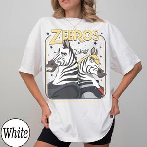 May include: White t-shirt featuring a cartoon graphic of two zebras with the text "ZEBROS" in yellow. The zebras are black and white with cartoonish expressions. The shirt has short sleeves and a crew neck.