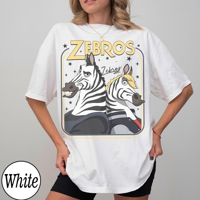 May include: White t-shirt featuring a cartoon graphic of two zebras with the text "ZEBROS" in yellow. The zebras are black and white with cartoonish expressions. The shirt has short sleeves and a crew neck.