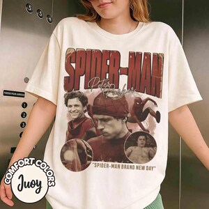 May include: Cream-colored t-shirt featuring a distressed graphic of Spider-Man with the text "SPIDER-MAN" and "Peter Parker." The design includes images of the character and the phrase "SPIDER-MAN BRAND NEW DAY." The shirt has short sleeves and a crew neck.
