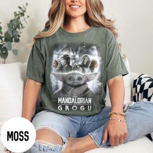 May include: Moss green t-shirt featuring a graphic of Grogu from The Mandalorian series. The design includes characters and the text "The Mandalorian and Grogu". The shirt is a relaxed fit.